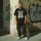 Homem veste t-shirt preta astredik com frase em branco, estilo hip-hop, em ambiente urbano com graffiti.