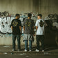 Três homens de costas vestindo t-shirts astredik com estampa graffiti amarela, em frente a uma parede grafitada.